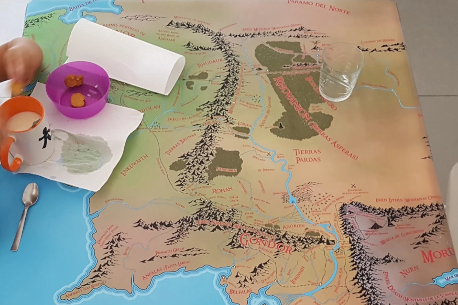 Un mantel de hule con el mapa de la Tierra Media de Tolkien impreso. Sobre él, un vaso de cristal, una taza con leche, unas servilletas de papel, un pequeño bol morado con dos galletas y una cucharilla.